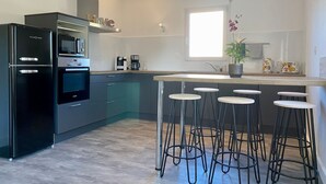 A modern kitchen with a built-in oven, microwave, and a coffee maker. There are bar stools and a window providing natural light.