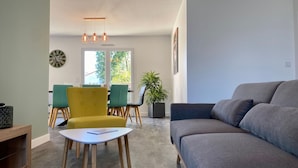 A modern living room with a grey sofa, a yellow armchair, and a white table. There is a dining area with green chairs and a table in the background.