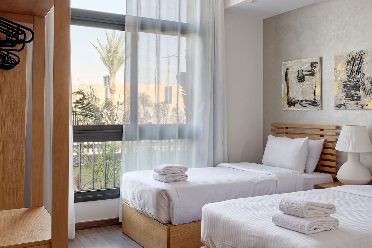 A modern hotel room with two beds, a wooden wardrobe, a large window with sheer curtains, and artwork on the wall.