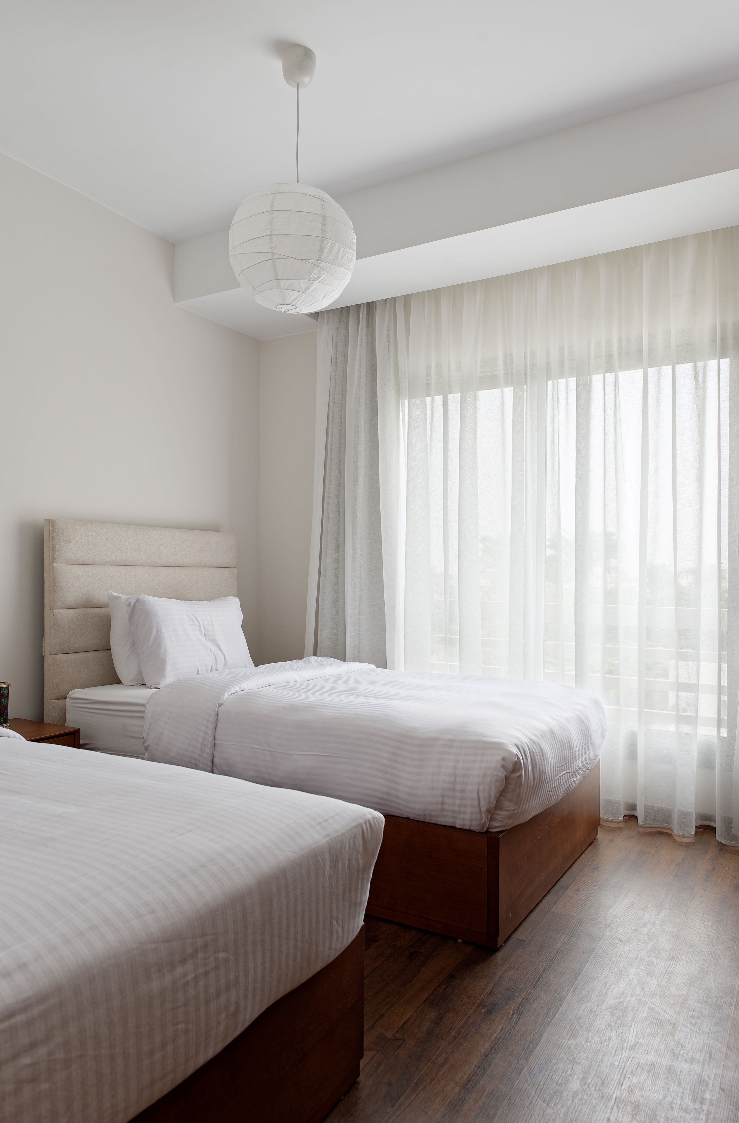 A hotel room with two beds, a large window with sheer curtains, and a hanging light fixture.