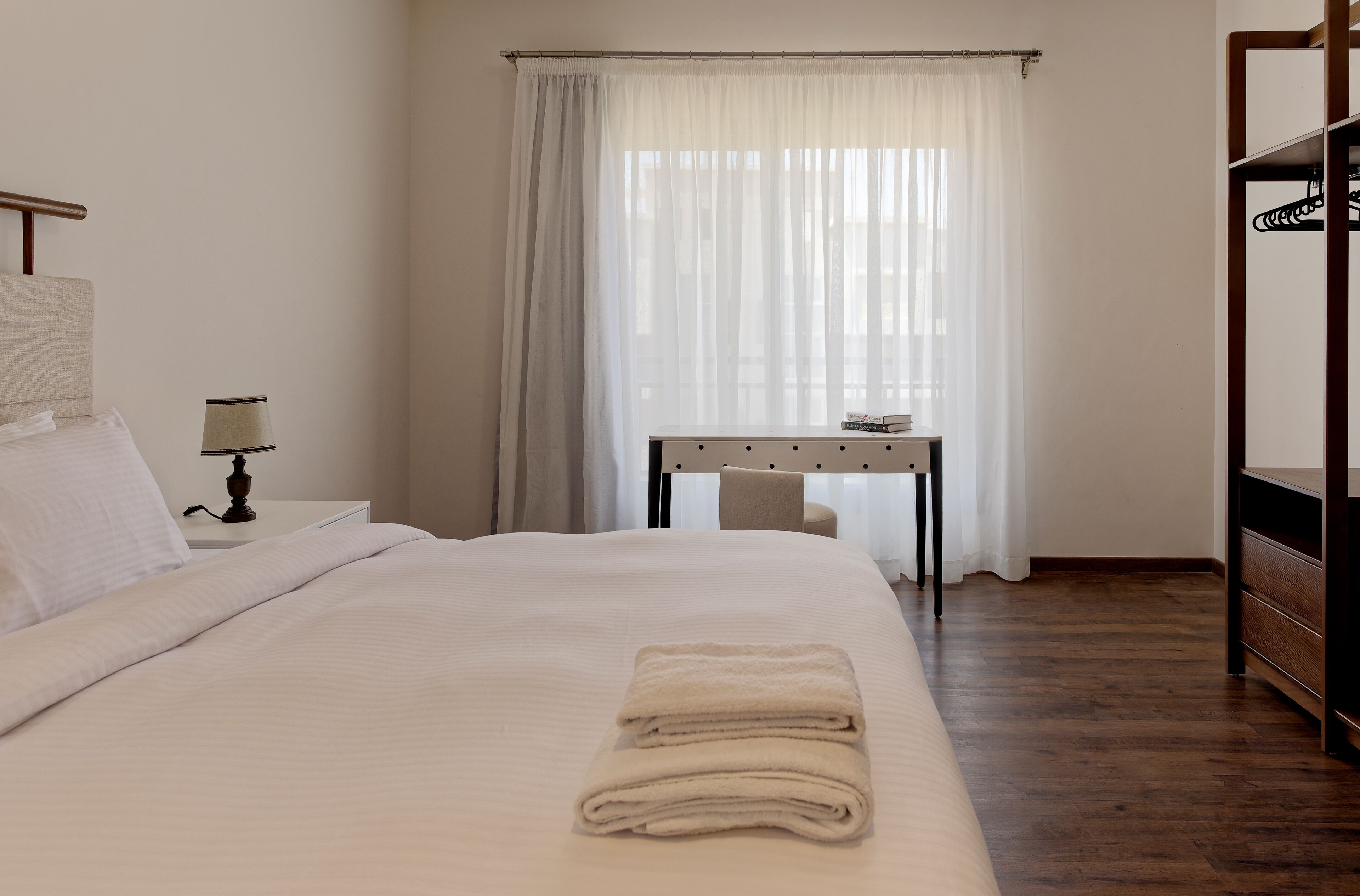 A neatly made bed with a folded towel, a bedside table with a lamp, a desk with a chair, and a wardrobe.