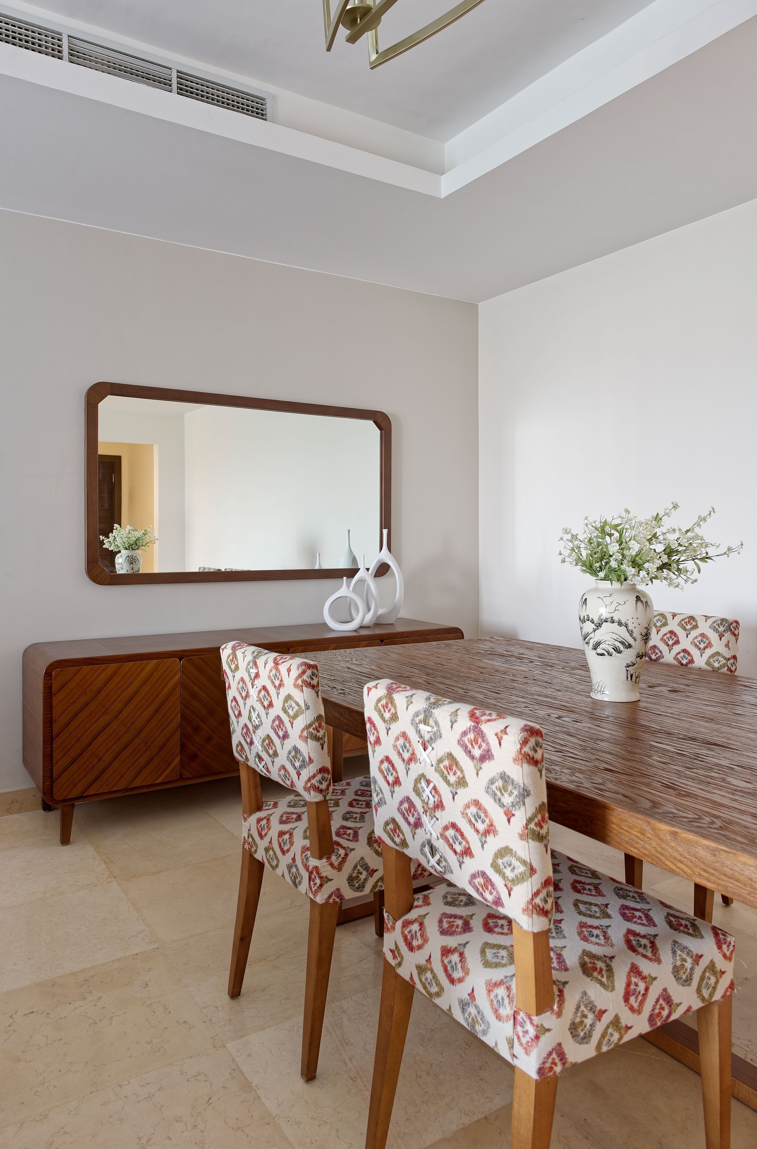 A dining area with a wooden table, patterned chairs, a vase with flowers, and a large mirror on the wall.