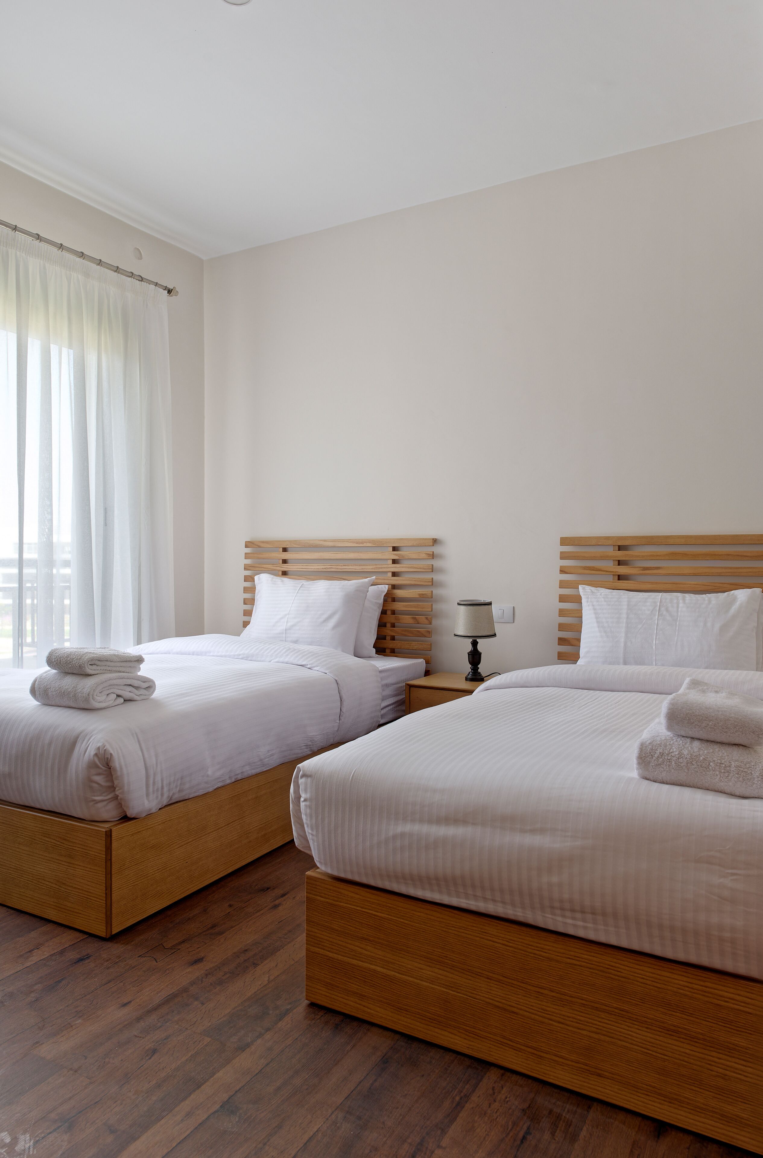 Une chambre d’hôtel avec deux lits, des têtes de lit en bois, du linge de lit blanc, une table de chevet avec une lampe et une fenêtre avec des rideaux légers.