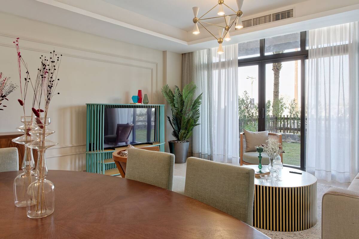 A modern living room with a wooden dining table, comfortable seating, a television, and a large window with curtains.