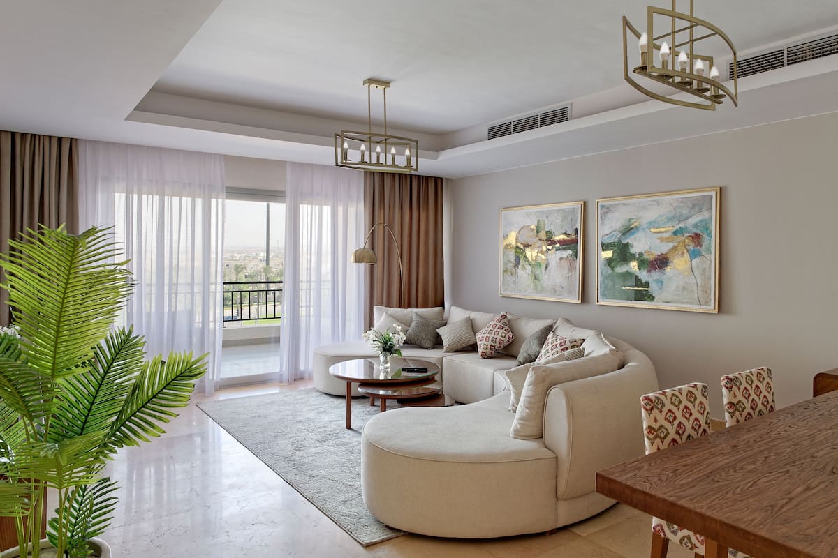 A modern living room with a sectional sofa, a round coffee table, and a dining table with chairs. There are two abstract paintings on the wall and a large potted plant.