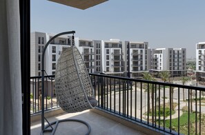 A balcony with a hanging chair, a view of modern apartment buildings, and a landscaped area with palm trees. - VESTA - Cairo Festival City Residence (New Cairo)