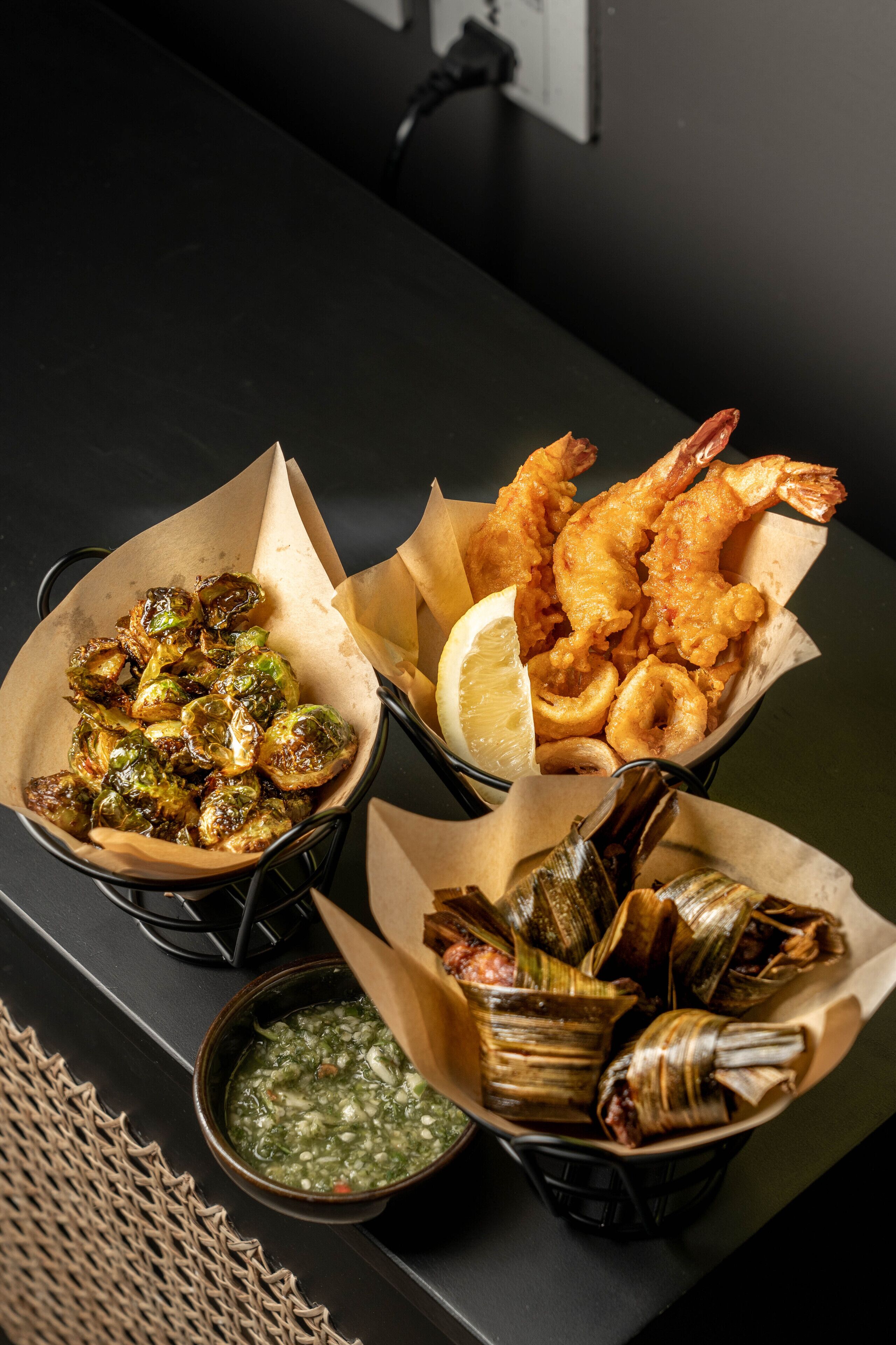 Uma mesa com três cestos de comida: brócolis de Bruxelas assados, camarões fritos e tamales envoltos em palha de milho.