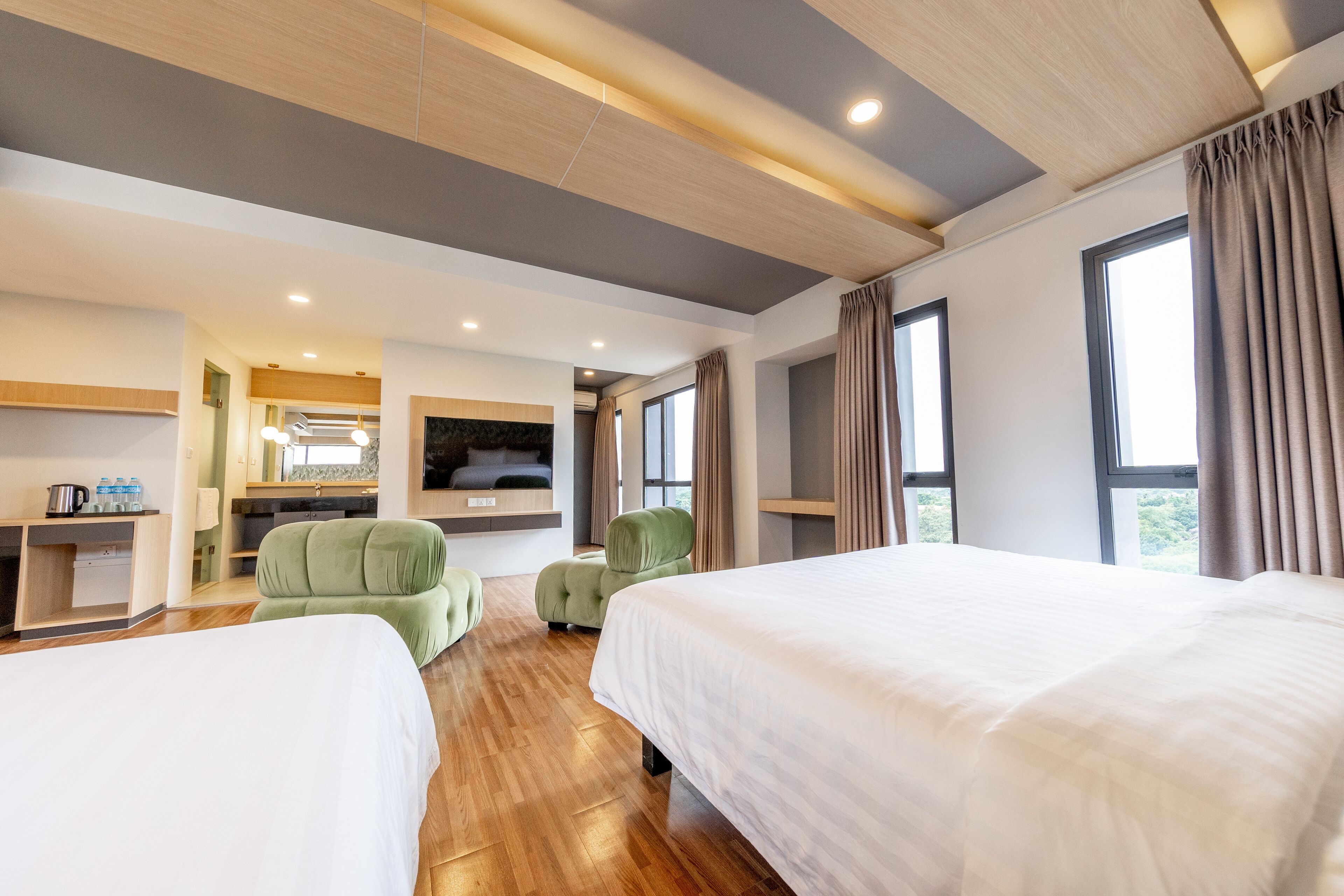Habitación de hotel con dos camas, una zona de estar con sillas verdes, una kitchenette y ventanas amplias.