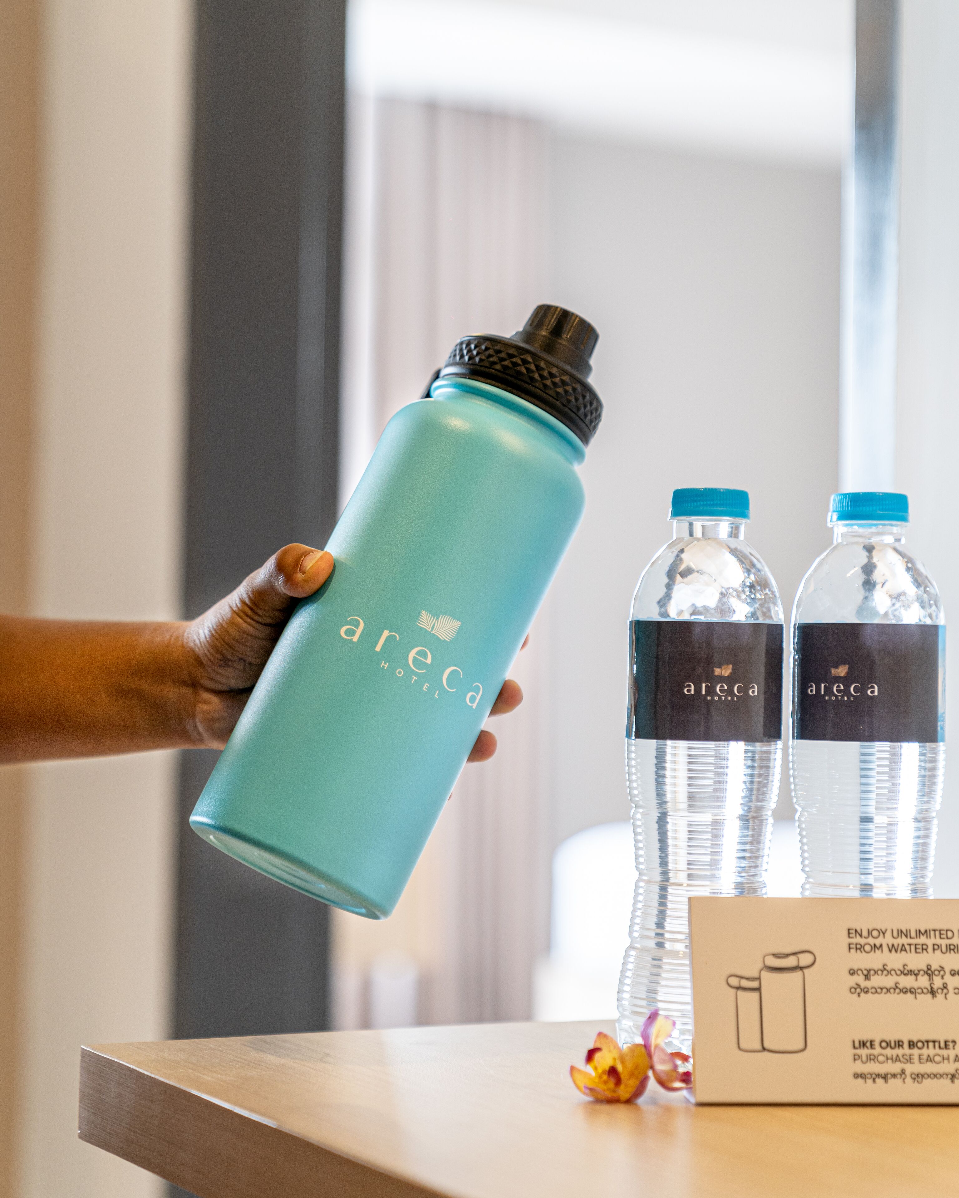 Una mano sosteniendo una botella de agua azul verdoso con tapón negro, y dos botellas de agua transparentes con tapones azules sobre una mesa.
