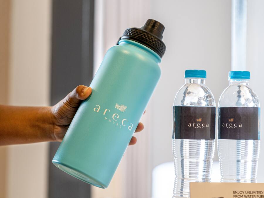 Una mano sosteniendo una botella de agua azul verdoso con tapón negro, y dos botellas de agua transparentes con tapones azules sobre una mesa.