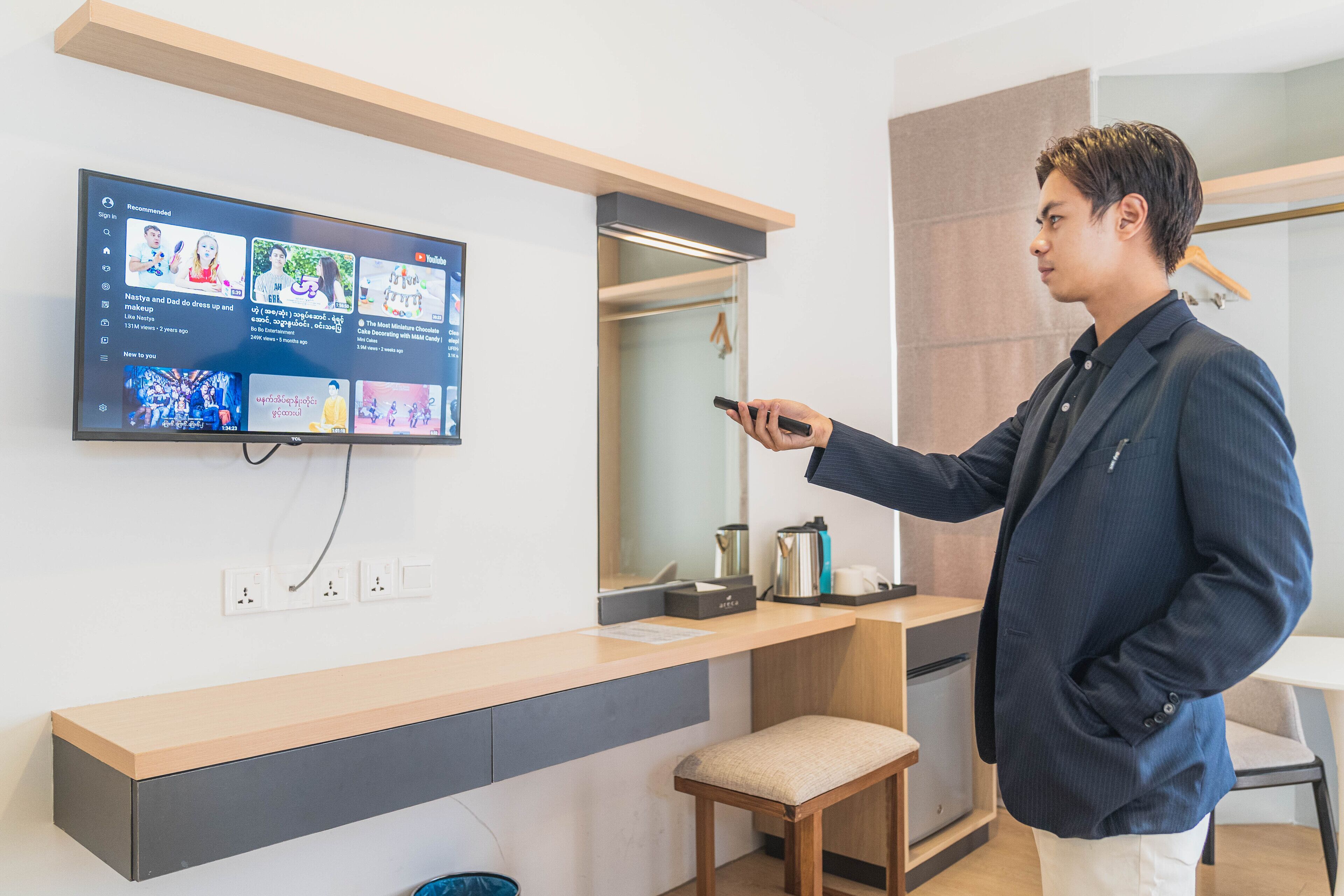 A modern hotel room with a flat-screen TV mounted on the wall displaying various content. There is a wooden counter with a bench, a small table with a kettle, and a hanging coat rack.