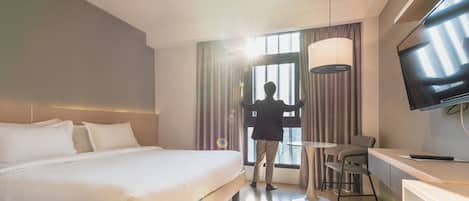 Habitación de hotel con cama, escritorio, silla, televisor y ventana con cortinas.