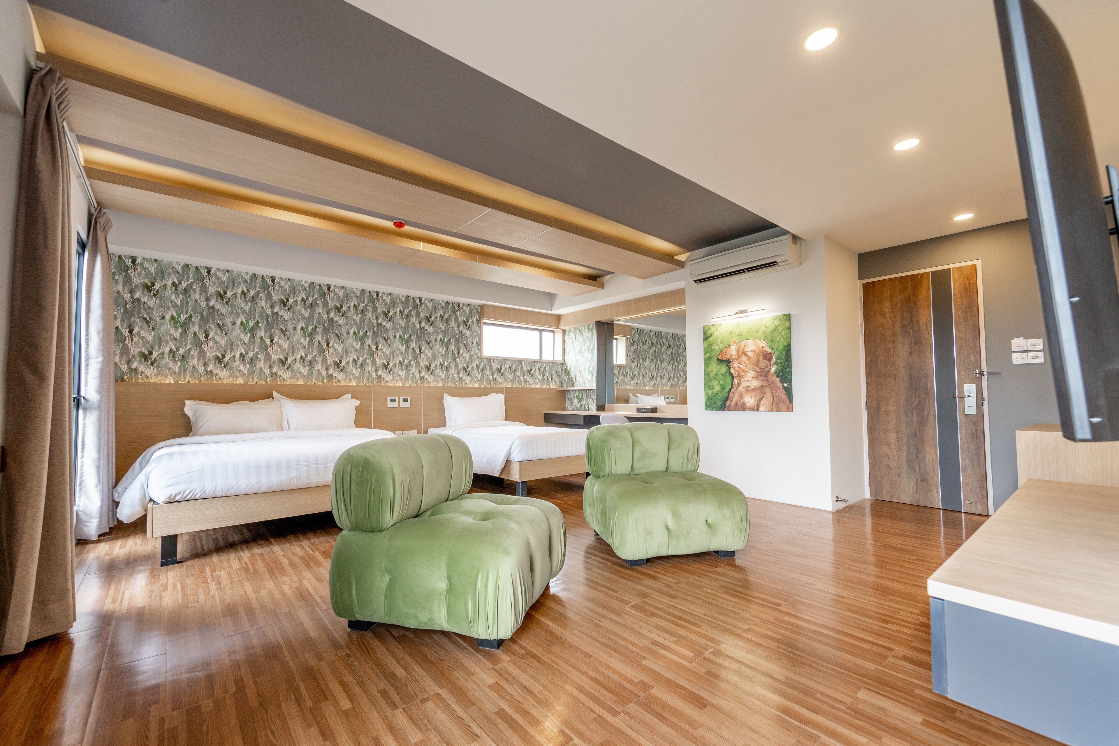 A modern hotel room with a wooden floor, a large bed, two green armchairs, and a desk.