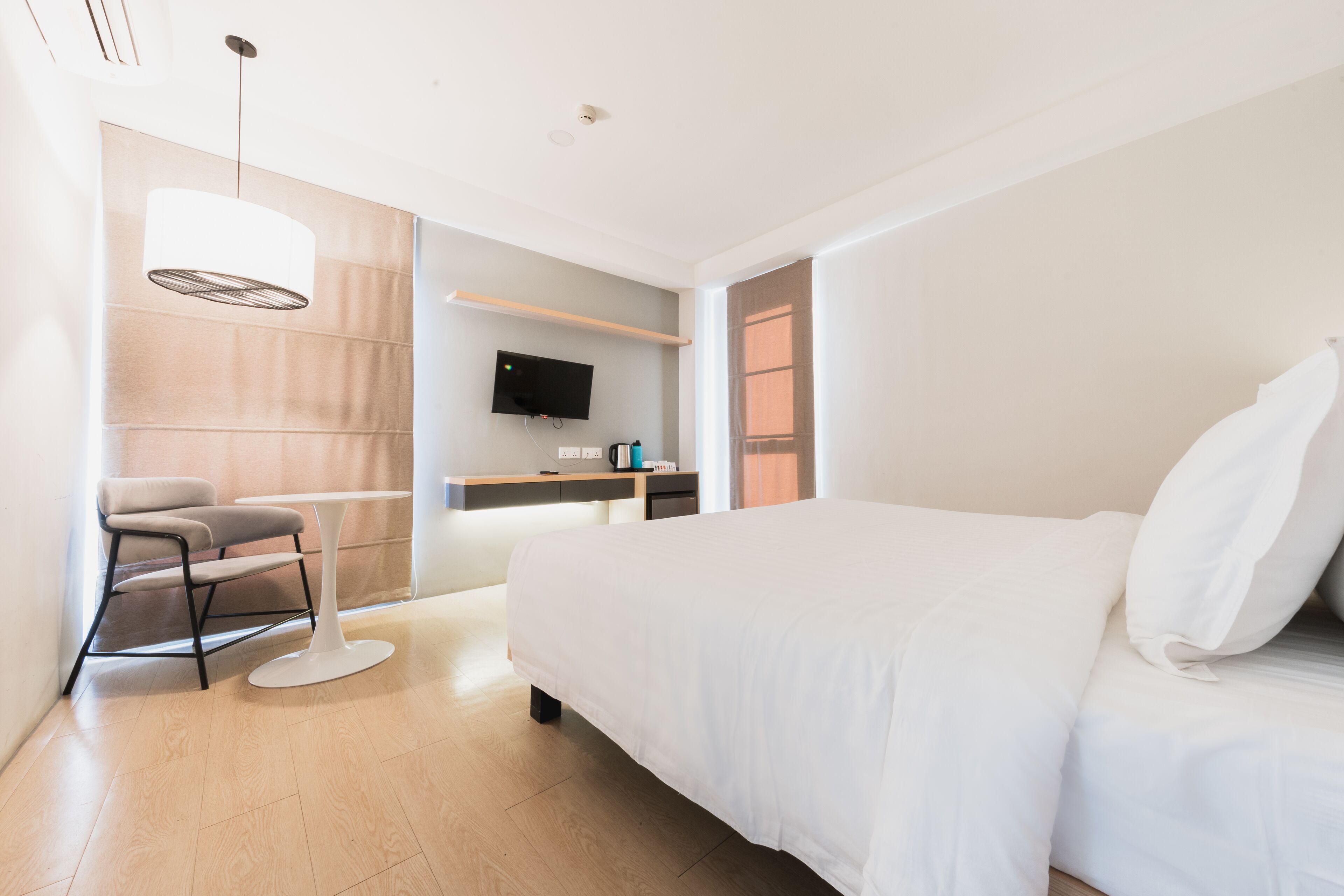 A modern hotel room with a large bed, a desk with a flat-screen TV, a chair, and a hanging light fixture.
