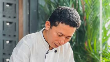 Un chef con uniforme blanco preparando comida en una cocina con un sofá rojo y una planta al fondo.