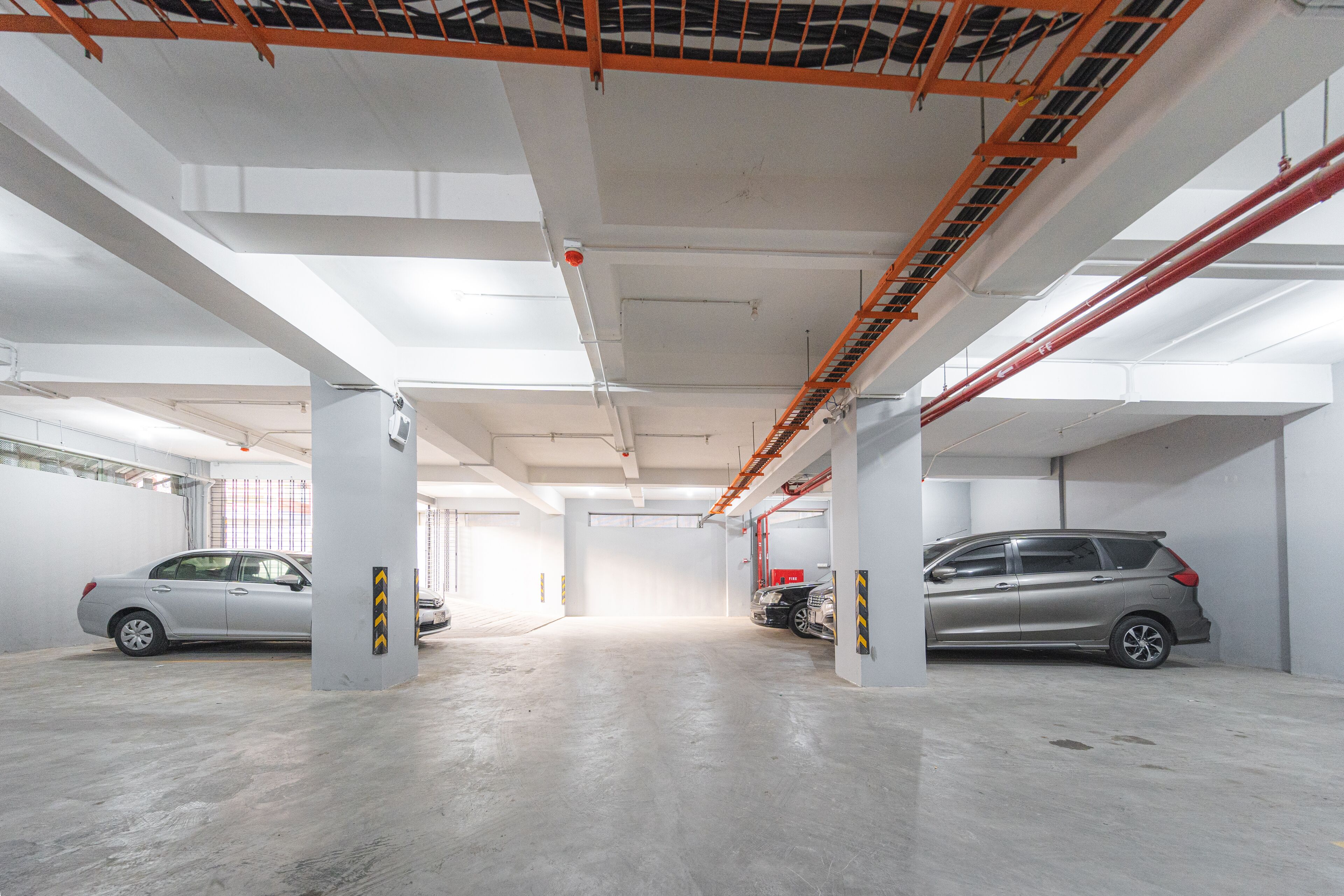 Un aparcamiento de varios niveles con suelos de hormigón y paredes blancas, que muestra dos coches estacionados y elementos estructurales como vigas y tuberías.