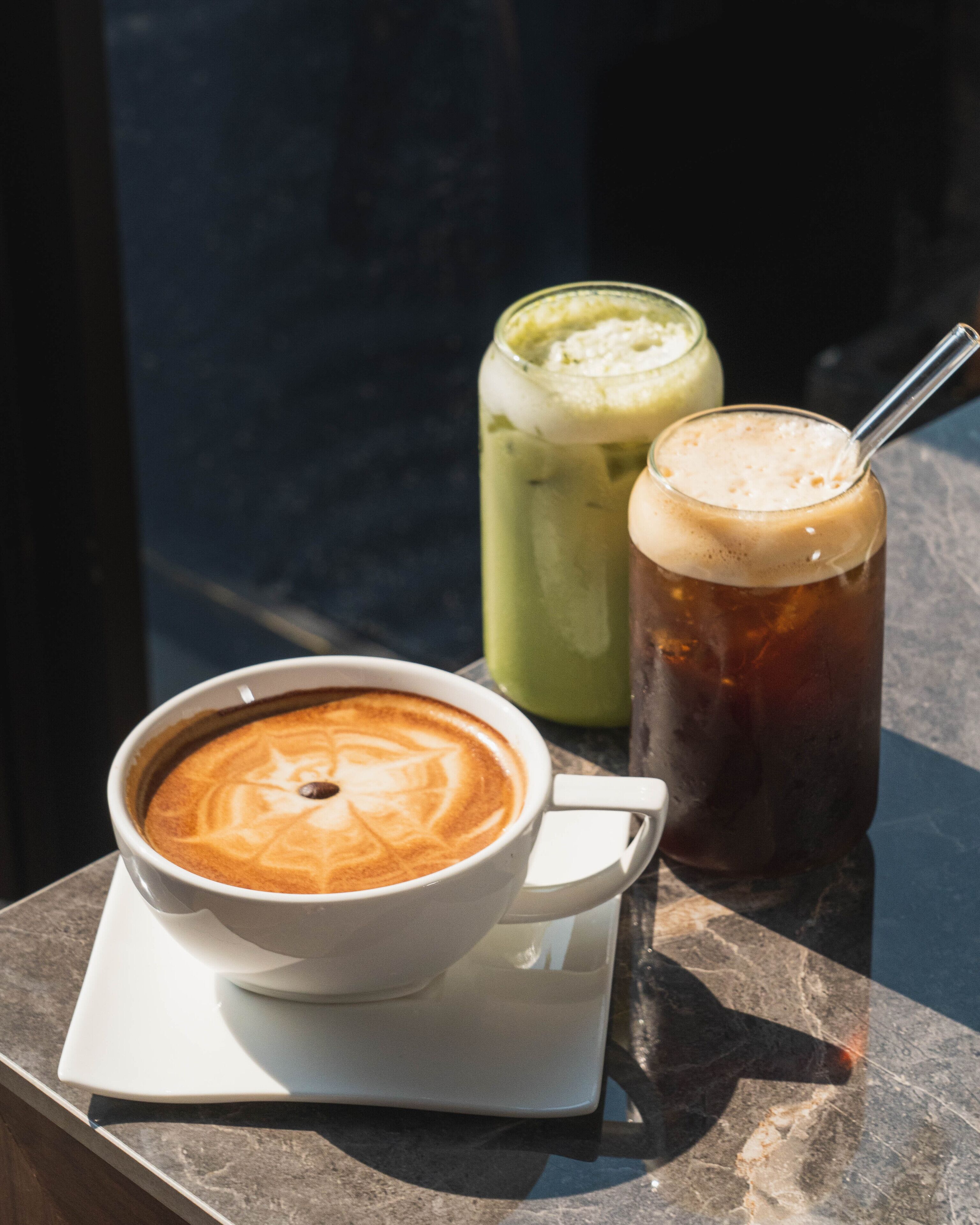 A cup of coffee with latte art on a white saucer, a glass of iced tea with a straw, and a green smoothie in the background.