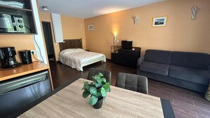 Habitació d'hotel senzilla amb llit, sofà, taula de menjador amb una planta, màquina de cafè i televisió.