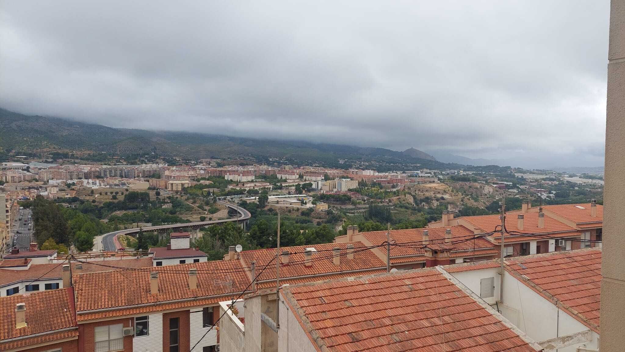 A cityscape with a winding road, buildings with red-tiled roofs, and a mountainous backdrop.