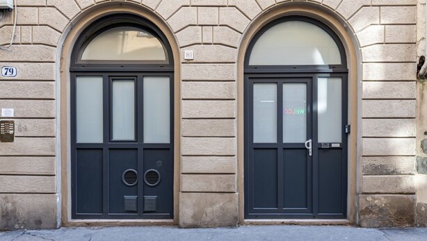 Eine Gebäudefassade mit zwei Rundbogenportalen, deren Nummern 79 und 81 sind, versehen mit satinierten Glasscheiben und einer Metall-Türklinke.