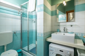 A bathroom with a glass shower enclosure, a toilet, a washing machine, and a sink with a mirror above it.