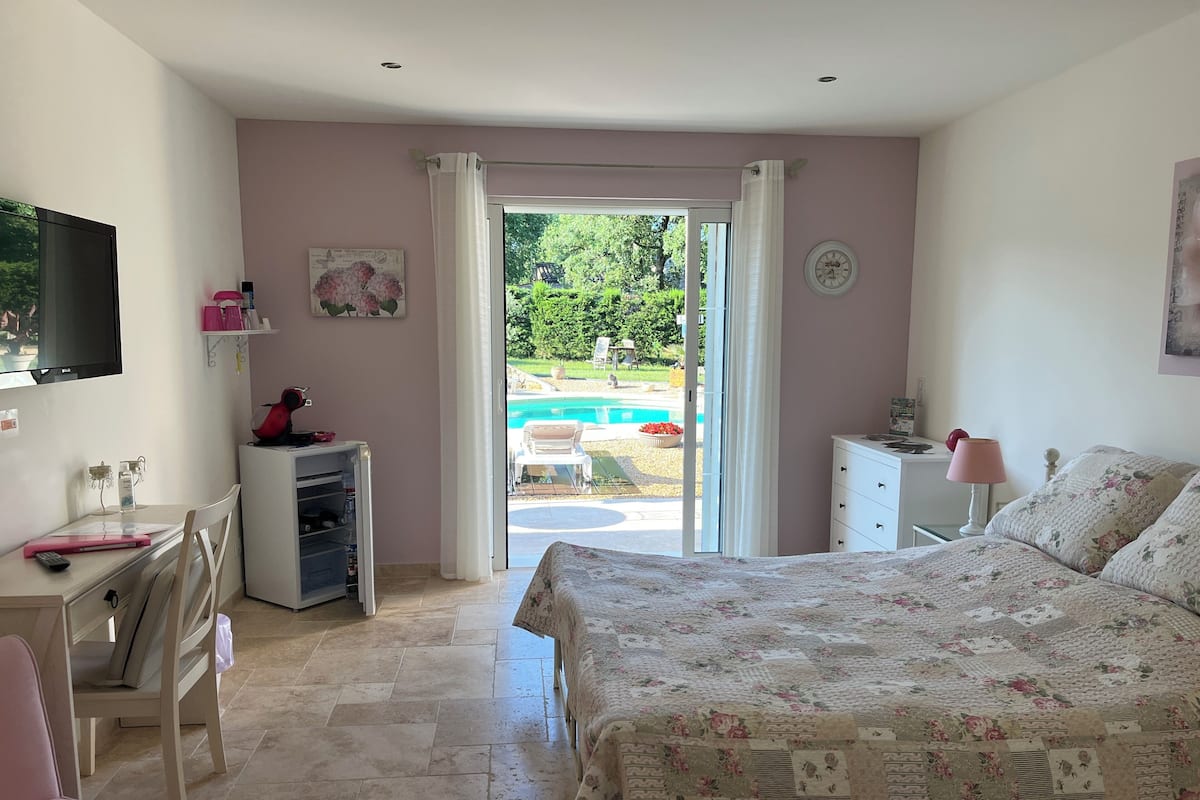 A bedroom with a bed, a desk, a chair, a TV, a small fridge, and a view of a pool outside.