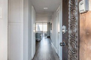 A hallway with white walls, a wooden door with a brass handle, and a view into a room with a patterned rug and a chair.