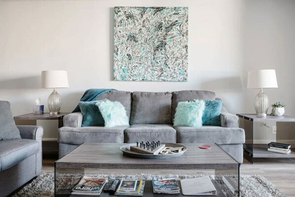 Un salon moderne avec un canapé gris, deux tables d’appoint avec des lampes, une table basse avec des magazines et un grand tableau abstrait accroché au mur.