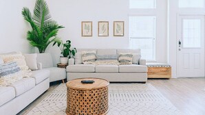 Una sala de estar moderna con un sofá gris claro, una mesa de centro redonda tejida y una planta grande en maceta. Hay tres obras de arte enmarcadas en la pared y una puerta blanca al fondo.