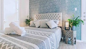 Una cama bien tendida con una colcha estampada y almohadas, un cabecero capitonado y una mesita de noche con una planta.