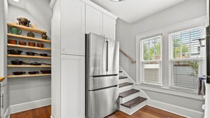 A compact kitchen with white cabinets, a stainless steel refrigerator, wooden shelves with cookware, and a window with blinds.