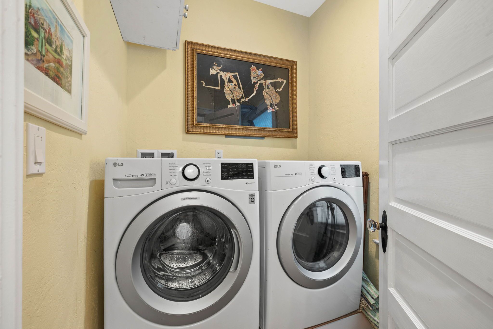 A laundry area with two LG front-loading washing machines, a framed artwork above them, and a white door to the right.