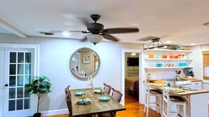 A dining area with a wooden table set for four, a ceiling fan, and a kitchen with white cabinets and a herringbone backsplash. - Downtown Cottage | Walk Everywhere, Park once (St Petersburg)