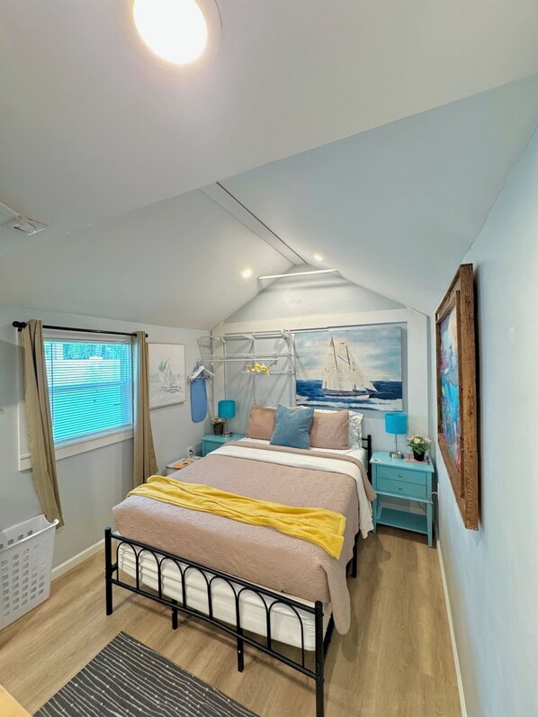 A bedroom with a bed, a window with curtains, a painting, and a nightstand with a lamp.
