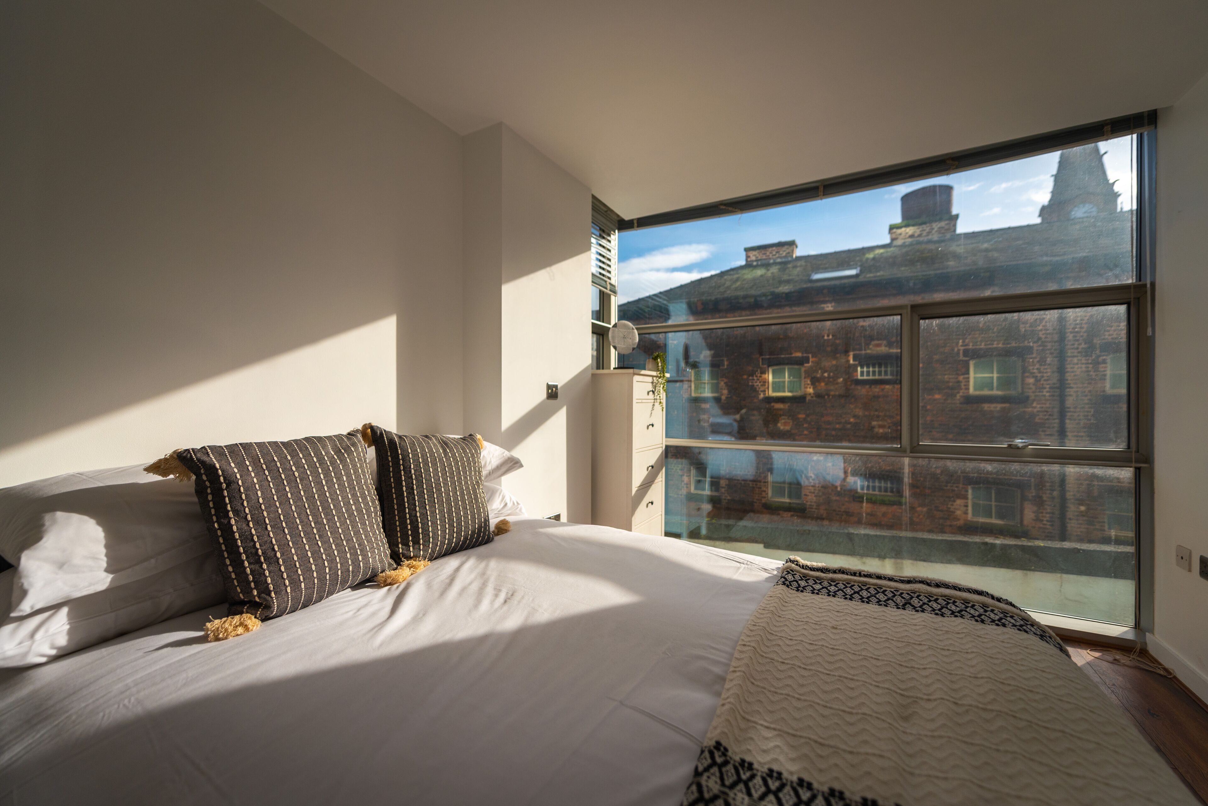 Ein Schlafzimmer mit großem Fenster, einem Bett mit weißer Bettwäsche und Blick auf Gebäude.