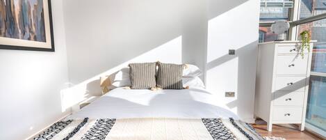 A modern bedroom with a large bed, a patterned rug, a white dresser, and a cityscape view.