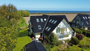 Ein Wohngebiet mit mehreren Häusern, die schräge Dächer und große Fenster haben, umgeben von üppigem Grün und Blick aufs Meer.