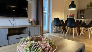 Ein modernes Wohnzimmer mit einem Holzesstisch, schwarzen Stühlen, einem an der Wand montierten Fernseher und einem großen Fenster.
