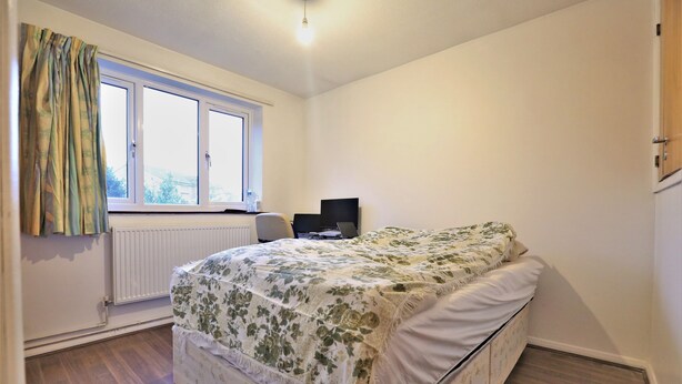 A bedroom with a bed, a window with curtains, a radiator, and a desk with a computer.