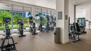 Ein modernes Fitnessstudio mit Cardiogeräten, Kraftmaschinen und Blick auf Palmen und ein Gebäude.