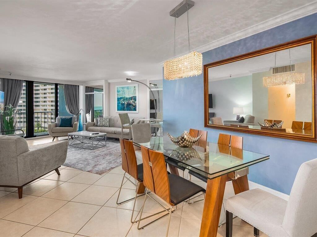 A modern living room with a glass dining table, comfortable seating, a large mirror, and a view of the outdoors.