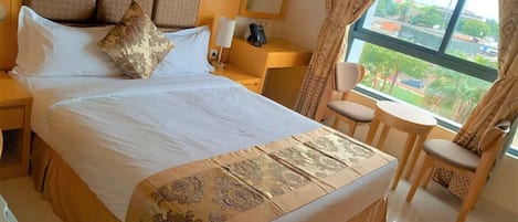 Habitación de hotel con una cama grande, una mesita de noche con lámpara, una silla, una pequeña mesa y vistas a la ciudad a través de una ventana.