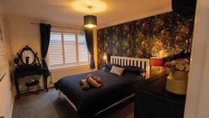 Een slaapkamer met een bed, nachtkastje, lamp, spiegel en raam met gordijnen.