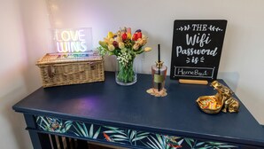 Een donkerblauwe tafel met een rieten mand, een glazen vaas met bloemen, een parfumflesje, een bordje met de tekst 'THE Wifi wachtwoord is HerneBay1', en een gouden olifantje.