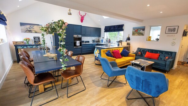 Een moderne keuken met donkerblauwe kastjes, een eettafel met bruine stoelen en een blauwe bank met rode kussens.