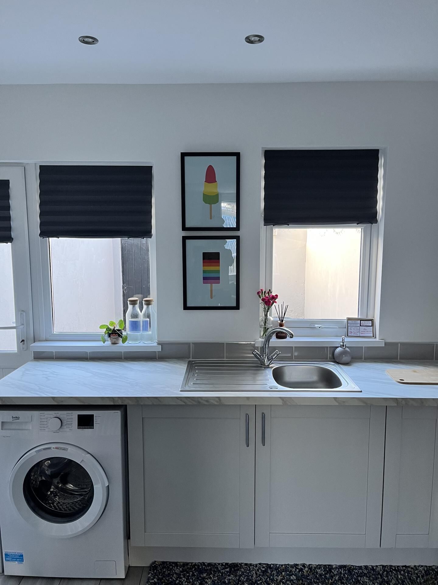 A modern kitchen with a built-in washing machine, a sink, and two windows with black blinds.
