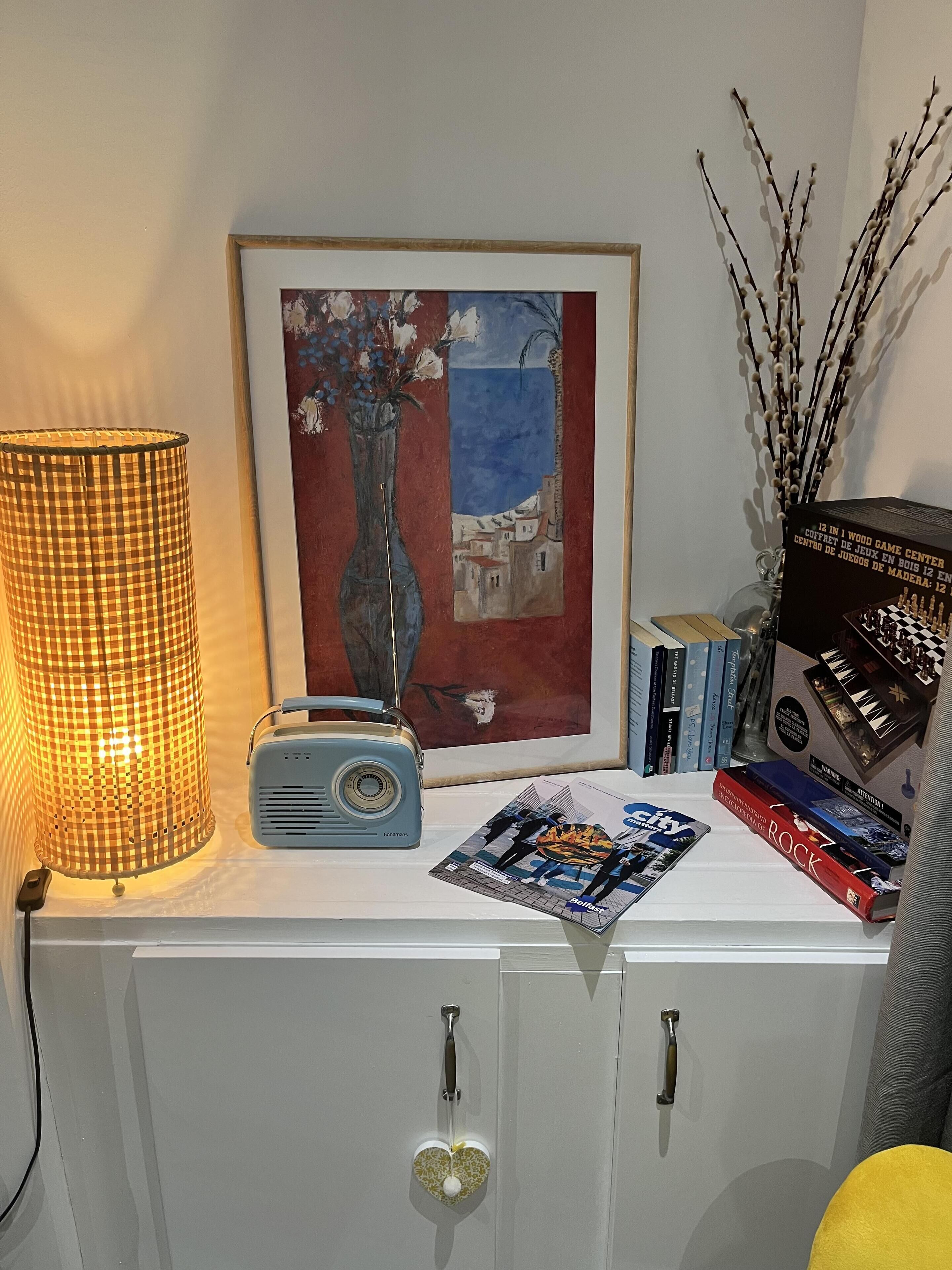 A room with a white cabinet, a framed painting, a radio, books, and decorative branches.