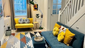 Una sala moderna con un sofá amarillo, un sofá azul y una alfombra estampada. Se ve una escalera a la derecha y una ventana con cortinas.