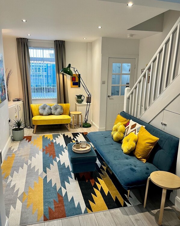 Uma sala de estar moderna com um sofá amarelo, um sofá azul e um tapete estampado. Há uma escada à direita e uma janela com cortinas.
