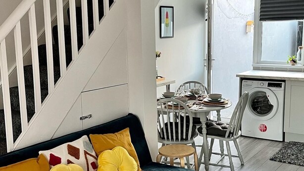 Una sala moderna con un sofá azul oscuro, cojines amarillos, una mesita redonda y una lavadora al fondo.